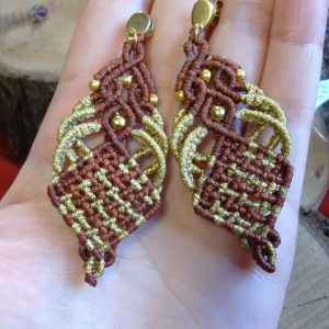 Ζευγάρι χειροποίητα μακραμέ σκουλαρίκια σε καφέ και χρυσό, με χρυσές χάντρες και ατσάλινα κουμπώματα, τοποθετημένα σε χέρι για παρουσίαση μεγέθους.