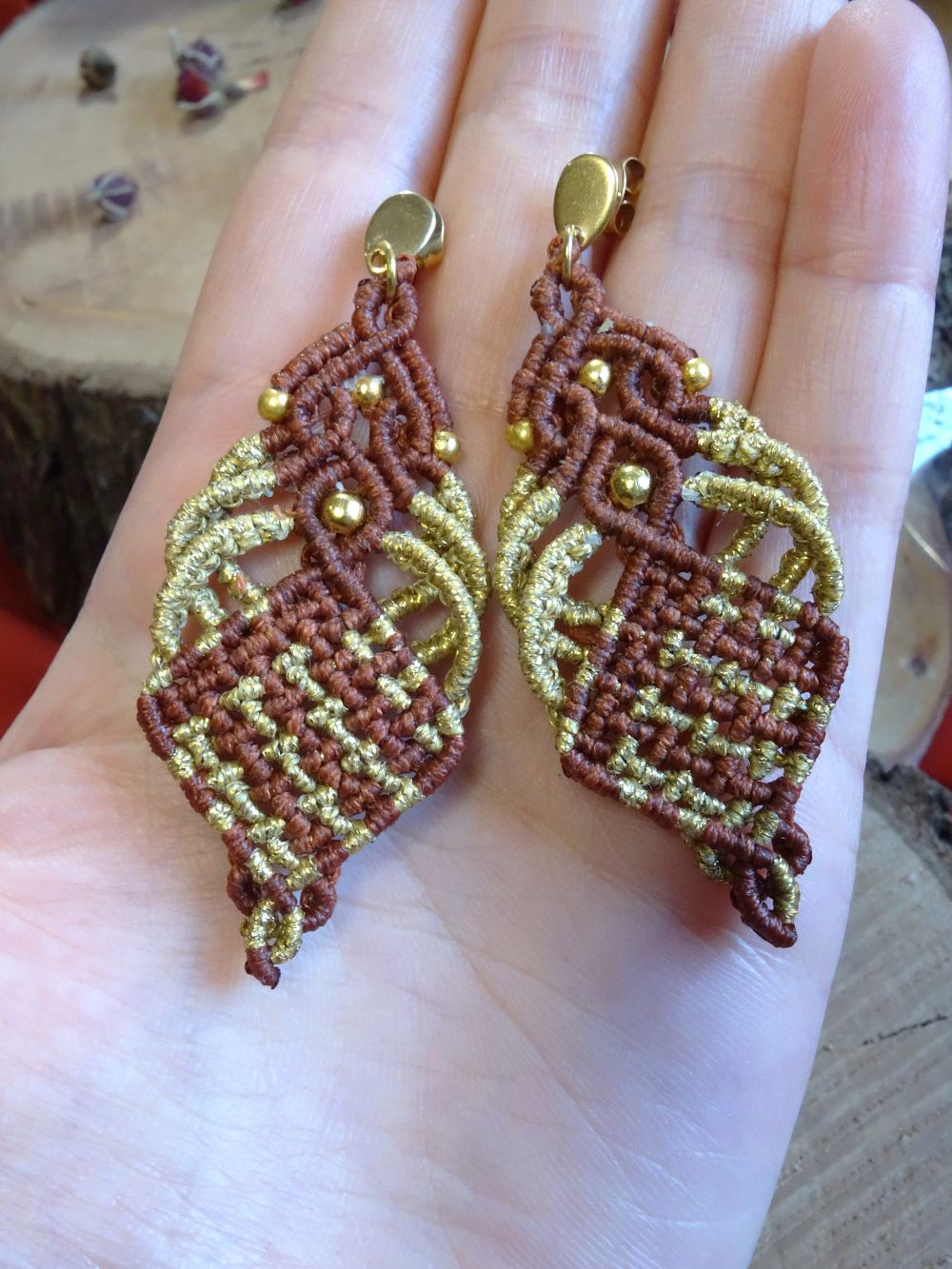Ζευγάρι χειροποίητα μακραμέ σκουλαρίκια σε καφέ και χρυσό, με χρυσές χάντρες και ατσάλινα κουμπώματα, τοποθετημένα σε χέρι για παρουσίαση μεγέθους.