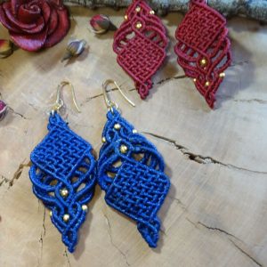 Χειροποίητα μακραμέ σκουλαρίκια σε γεωμετρικό σχέδιο, ένα ζευγάρι σε κόκκινο χρώμα και ένα σε μπλε, διακοσμημένα με χρυσές χάντρες, τοποθετημένα πάνω σε ξύλινη επιφάνεια με φυσικά στοιχεία.