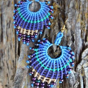 Χειροποίητα μακραμέ σκουλαρίκια σε σχήμα βεντάλιας, σε αποχρώσεις μπλε, πράσινου και μπεζ, διακοσμημένα με μωβ χάντρες, τοποθετημένα πάνω σε φυσικό ξύλο.