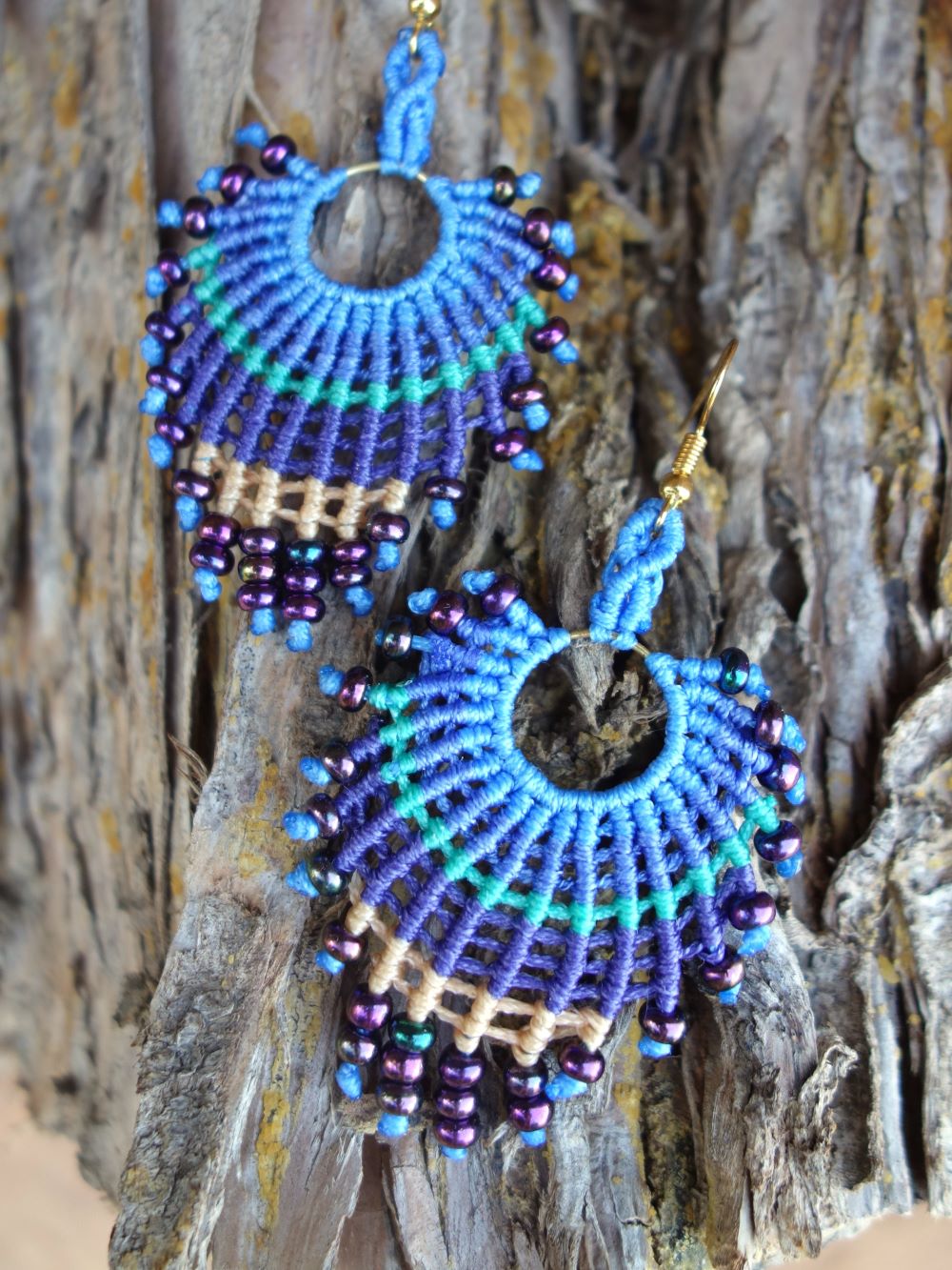 Χειροποίητα μακραμέ σκουλαρίκια σε σχήμα βεντάλιας, σε αποχρώσεις μπλε, πράσινου και μπεζ, διακοσμημένα με μωβ χάντρες, τοποθετημένα πάνω σε φυσικό ξύλο.