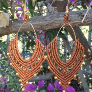 Χειροποίητα μακραμε σκουλαρίκια σε καφέ χρώμα με χρυσές χάντρες, κρεμασμένα σε κλαδί με φόντο μωβ λουλούδια