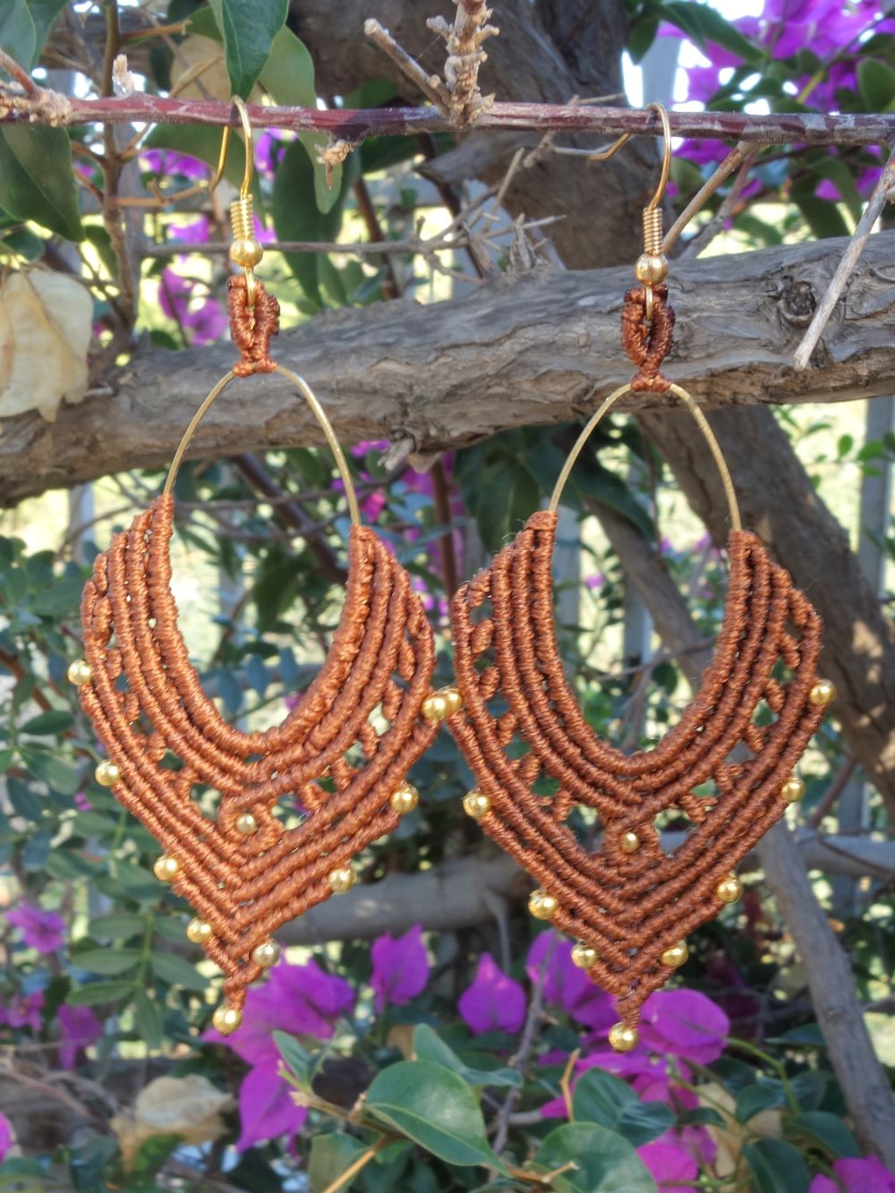 Χειροποίητα μακραμε σκουλαρίκια σε καφέ χρώμα με χρυσές χάντρες, κρεμασμένα σε κλαδί με φόντο μωβ λουλούδια
