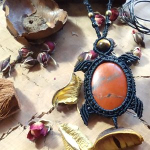 Η φωτογραφία δείχνει το κολιέ πάνω σε ξύλινο φόντο, με φυσικά στοιχεία όπως ξερά άνθη και βότανα γύρω του, αναδεικνύοντας τον boho-φυσικό χαρακτήρα του. Το κόκκινο του ίασπη λάμπει στο κέντρο, ενώ η μαύρη πλέξη σε σχήμα χελώνας τον αγκαλιάζει αρμονικά.