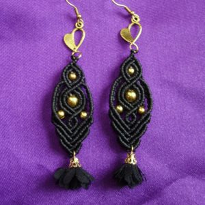 Ζευγάρι χειροποίητα μακραμέ σκουλαρίκια σε μαύρο με χρυσές χάντρες και φουντίτσα, τοποθετημένα σε μωβ φόντο