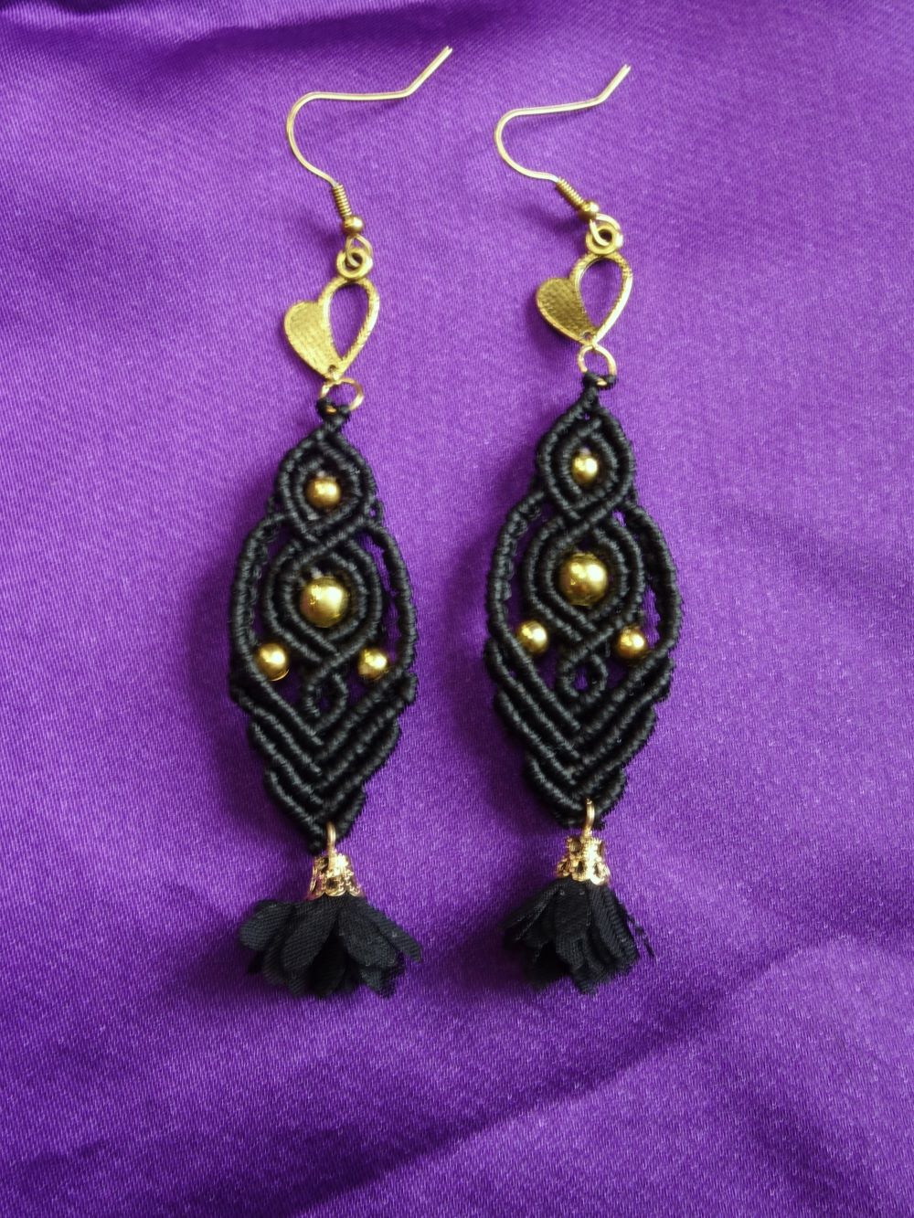 Ζευγάρι χειροποίητα μακραμέ σκουλαρίκια σε μαύρο με χρυσές χάντρες και φουντίτσα, τοποθετημένα σε μωβ φόντο