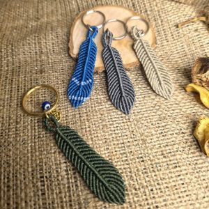 Χειροποίητα μπρελόκ μακραμέ σε σχήμα φτερού σε μπλε, ανθρακί, μπεζ και πράσινη απόχρωση, τοποθετημένα πάνω σε λινό ύφασμα.