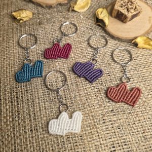 Χειροποίητα μπρελόκ μακραμέ σε σχήμα διπλής καρδιάς σε διάφορα χρώματα, τοποθετημένα πάνω σε λινό ύφασμα.