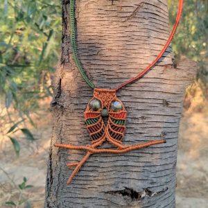 Χειροποίητο μακραμέ κολιέ σε σχήμα κουκουβάγιας με καφέ, πράσινα και κεραμιδί νήματα και χάντρες στα μάτια, πάνω σε φόντο δέντρου.