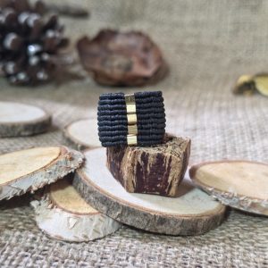 Μαύρο μακραμέ δαχτυλίδι με χρυσές ατσάλινες χάντρες επάνω σε ξύλινη βάση.