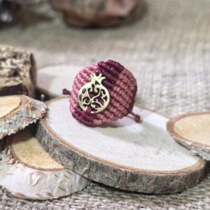 Μακραμέ δαχτυλίδι σε ροζ και μπορντό με χρυσό ατσάλινο ρόδι, τοποθετημένο επάνω σε ξύλινη βάση.