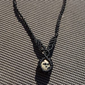 Μαύρο μακραμέ κολιέ με μικρή πέτρα Ιάσπη Δαλματίας πάνω σε υφασμάτινο γκρι φόντο.