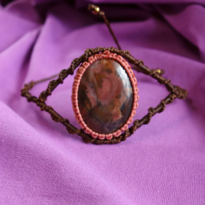 Μακραμέ βραχιόλι με οβάλ πέτρα Ιάσπη σε καφέ και ροζ αποχρώσεις πάνω σε μοβ υφασμάτινο φόντο.