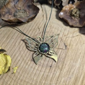 Χειροποίητο ορειχάλκινο κολιέ πεταλούδα με διατρητό σχέδιο και ημιπολύτιμο ρουμπίνι ζωϊσίτη πάνω σε ξύλινη επιφάνεια