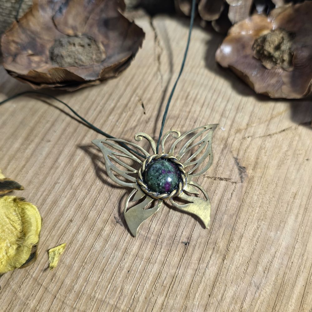 Χειροποίητο ορειχάλκινο κολιέ πεταλούδα με διατρητό σχέδιο και ημιπολύτιμο ρουμπίνι ζωϊσίτη πάνω σε ξύλινη επιφάνεια