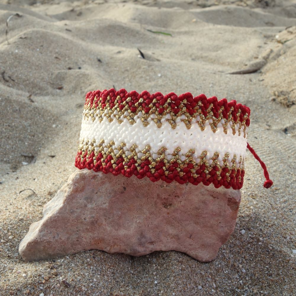 Χειροποίητο μακραμέ βραχιόλι σε κόκκινο λευκό και χρυσό με γεωμετρικό μοτίβο πάνω σε φυσικό φόντο άμμου